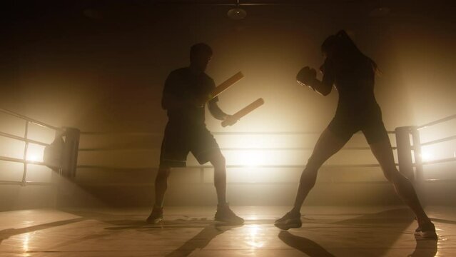 Close-up view of female kickboxer training with her coach at boxing club. Active woman having intensive workout in the gym. High quality 4k footage in golden orange yellow foggy light