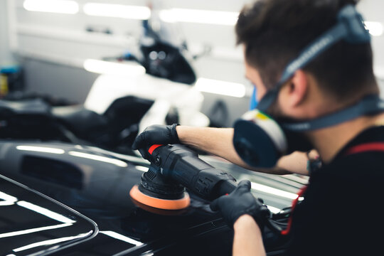 Rear view of white man in protective face mask polishing top of black car using professional buffer tool. Car detailing process. Blurred foreground. Horizontal indoor shot. High quality photo