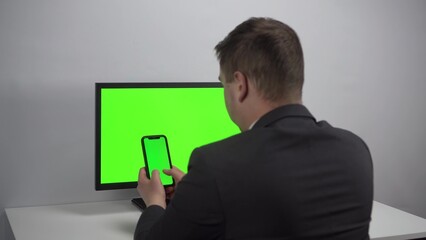 A young businessman sits at a computer and a phone in his hands with green screens. A man works in an office in a jacket. View from the back.