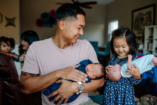 Father holding newborn, smiling at his toddler daughter in living room