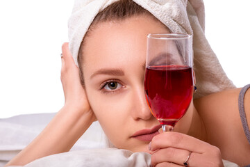 Beautiful young sexy caucasian girl with a white towel on her head holding a glass of wine and looking through it on a white isolated background shot in studio