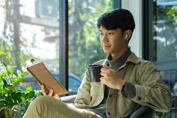 Handsome hipster man relaxing holding coffee cup and reading book while sitting on chair in cafe.