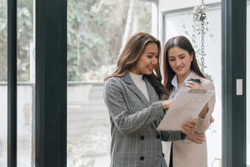 Two young pretty asia business woman in suit talking together in modern office workplace, Thai woman, southeast asian, Standing