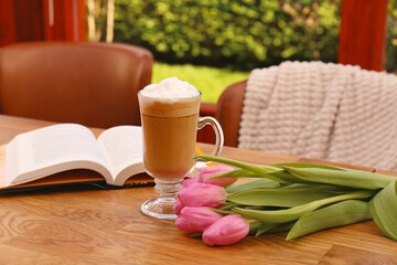 Glass of delicious cocoa, pink tulips and book on wooden table at terrace