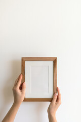 Woman's hands holding a wooden stylish blank frame against a white wall. Frame mock up, copy space...