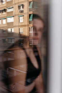 Non binary person standing near window with reflection of city