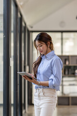 Young business Asian woman using tablet ipad, standing near the window in workplace