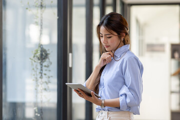 Young business Asian woman using tablet ipad, standing near the window in workplace