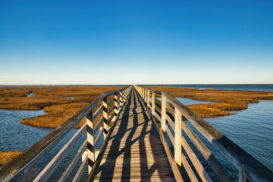 Under a clear blue Winter sky, an empty  boardwalk stretches toward an ocean horizon during high tide at Gray's Beach, near Yarmouth Port, MA, on Cape Cod.