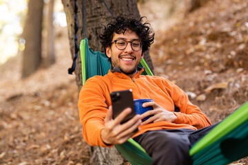 Adventurous Man Holds A Blue Mug And Uses Phone While In A Hammock