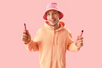 Young man in bucket hat with disposable electronic cigarettes on pink background