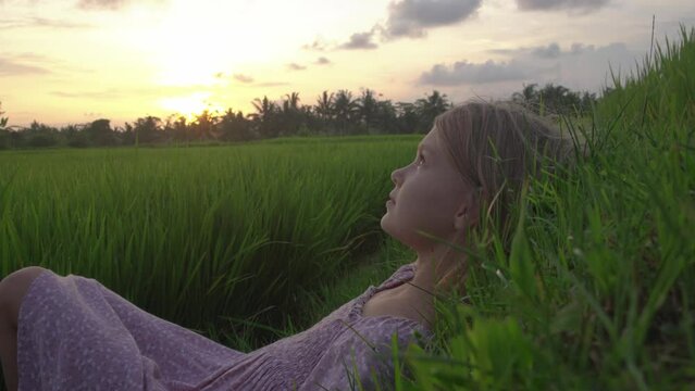The girl lies on the grass and enjoys a beautiful view of the green fields at sunset