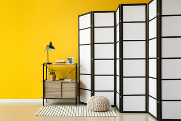 Interior of room with folding screen, shelving unit and pouf near yellow wall