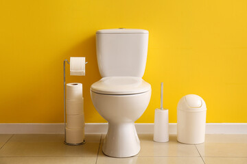 Ceramic toilet bowl, paper rolls, bin and brush near yellow wall