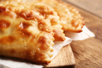 Cutting board with tasty chebureks and parchment on wooden background, closeup