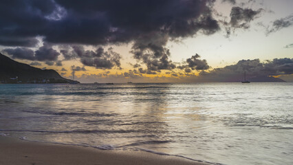 Twilight on the beach of a tropical island. Purple clouds in the sky, highlighted in orange.  Reflection on the ocean. Waves are spreading across the sand. Yachts and boats on the horizon. Seychelles