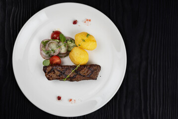 Grilled beef skirt steak meat with potato and vegetables on white plate on wooden black background