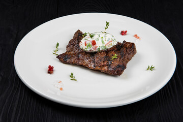 Grilled skirt beef meat steak on a plate with white vegetable sauce decorated by herbs and berries. Dark wooden background