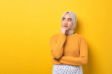 Beautiful pensive Asian girl wearing hijab looking aside at empty space, touching chin, thinking about offer isolated on yellow background