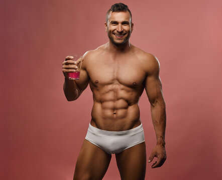 Muscular handsome man smiling and holding glass with liquid at pink background. Male fitness model posing in studio. Caucasian hunk with six pack abs.