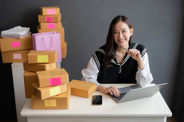 Asian women are online sellers. have a beautiful smile happy face Using a laptop to see a list of business orders from customers on the table. Around it are boxes, shopping bags, pens and smartphones.