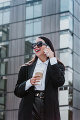 Young hispanic businesswoman in coat with coffee cup and talking on mobile phone  while intently looking away on city street in Mexico Latin America