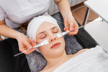 Woman getting facial peeling by cosmetologist. Beautician applying clay face mask on woman face. Cosmetologist applying mask on clients face in spa salon. Spa cosmetologist applying facial mask.