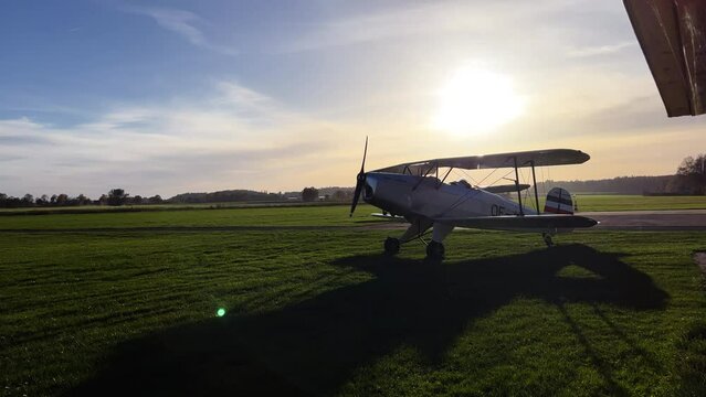 Medium wide shot of an old vintage airplane standing on a grass rollway getting ready. Starting propellers. Evening sun in the background.