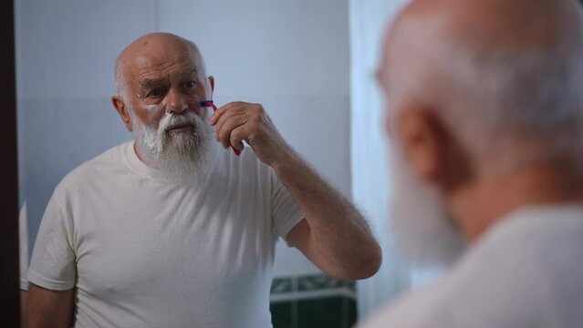 Reflection in mirror of senior Caucasian man shaving stubble on cheek with razor. Concentrated confident old retiree styling white beard at home in bathroom. Hygiene and lifestyle concept