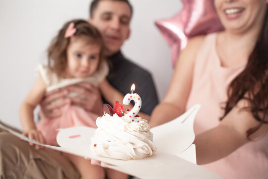 Happy Multinational Family, Asian Mother Coucasian Father And Daughter Celebrate The Birthday Of Two Years Old Kid. The Girl Blows Out Candle On The Cake Against Thr Background