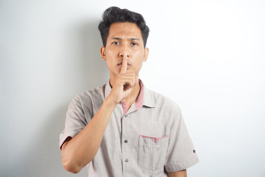 silent hand symbol or shhh (ssst). portrait of asian man glaring while looking at camera with index finger in front of mouth. Indonesian man wearing a hoodie on a white background