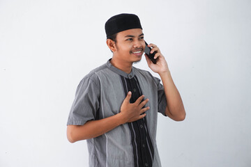 smiling young asian muslim man holding chest and holding phone calling by mobile phone wearing koko...