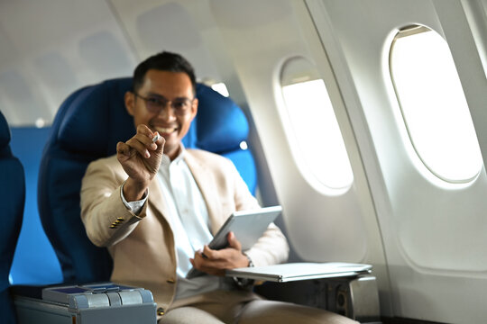 Image of smiling businessman in elegant luxury suit using digital tablet while sitting in airplane cabin during business travel