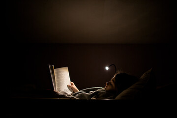 Young girl reading before bedtime in bed with lamp