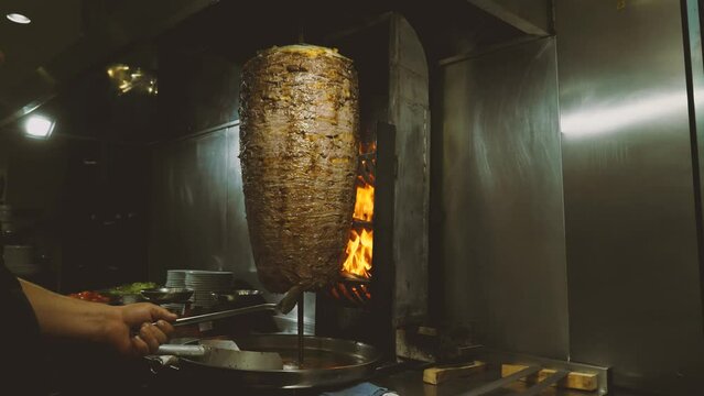 Doner kebab or shawarma on a rotating vertical spit. The chef spins the spit with a long metal spatula. Flames from the coals are burning. Fat drips from the meat. Traditional street fast food