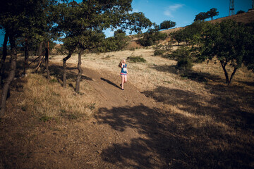 An attractive blonde girl in striped shorts and a blue t-shirt walks along a path in the hills, around the trees, on a bright sunny day.