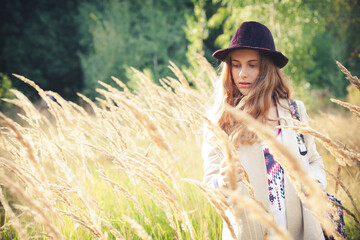 A stylish young girl with blond hair, in a hat and a light jacket, walks through a field with tall grass, on a bright sunny summer evening.