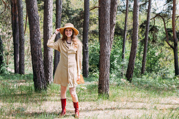 A slender, young girl in a beige sundress and red boots, stands in the forest against the backdrop of trees, on a bright sunny day. He holds a straw hat in his hands and smiles.