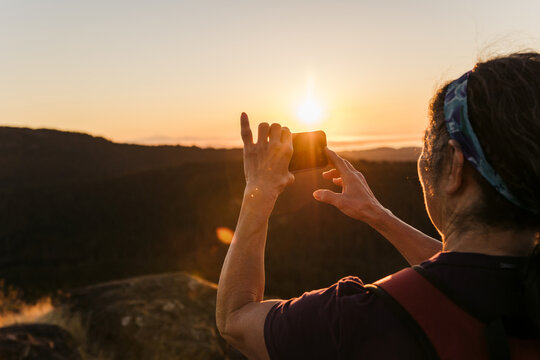 Woman taking picture with mobile phone at sunrise on hilltop.