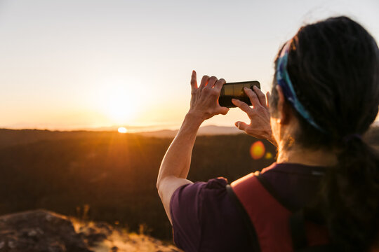 Woman taking picture with mobile phone at sunrise on hilltop.