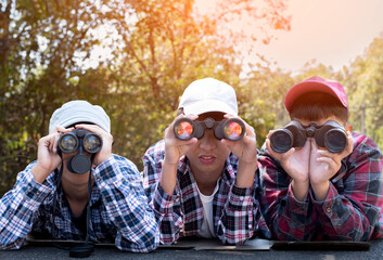 Asian boys are using binoculars to do the birds' watching in tropical forest during summer camp,...