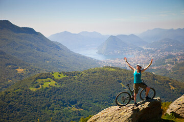 A female mountain biker is standing on top of a rock with her hands happy in the air.