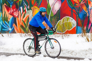 A bearded man rides a bicycle in a winter park against the background of a multi-colored wall. Eco-friendly transport in winter. Active lifestyle