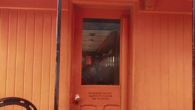Vintage orange passenger train revealed through handheld shot, tight close-up from exterior while in motion, swaying.