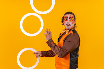 Portrait of a juggler in a vest with a painted face juggling hoops on a yellow background