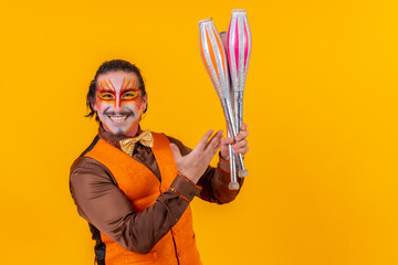 Portrait of a juggler in a vest with a painted face juggling with maces on a yellow background - obrazy, fototapety, plakaty