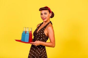 Smiling lady in polka dot dress and retro hairstyle holds a tray with bottles of soda in her hands...