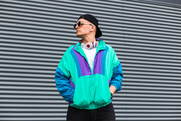 A young man in a sports jacket, cap, sunglasses and headphones around his neck in retro 90s style. Vintage look in modern fashion. The revival of old trends in a new way