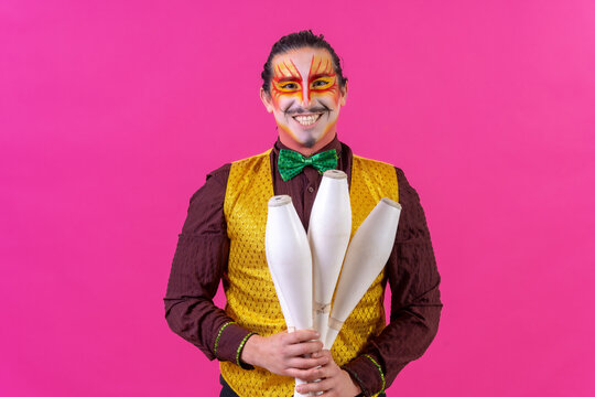 Juggler in a vest and with painted face juggling maces on a pink background