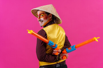 Portrait of a clown with white facial makeup on a pink background, with toy guns and a Chinese hat